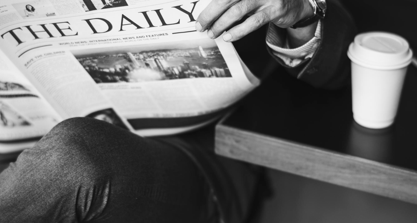 Black and white photo of a person reading a newspaper with a coffee cup on the table